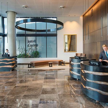 Pullman Brisbane Airport Hotel Exterior foto The image shows a modern hotel lobby. There are two reception desks made of dark-colored materials, and staff members are attending to guests at these desks. The lobby features large windows that allow natural light to fill the space, and there are p
